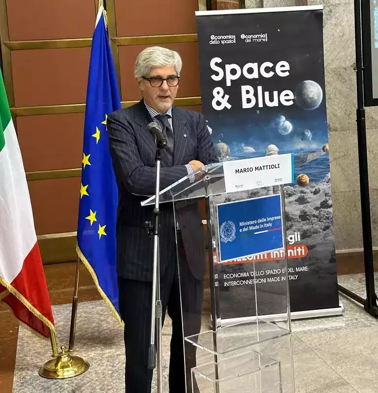 Ritratto di Mario Mattioli a un evento Space & Blue con bandiera italiana, europea e poster.